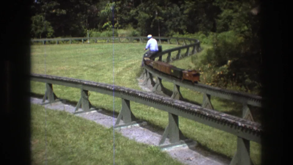 The miniature train curves along a raised outdoor railway in Baxter, Maine, with the rider seated near the front and trees enclosing the green yard.