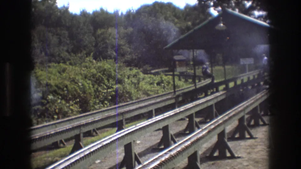 The elevated miniature railway runs toward a small green-roofed station shelter, with wooded vegetation and 8mm film marks visible in the frame.
