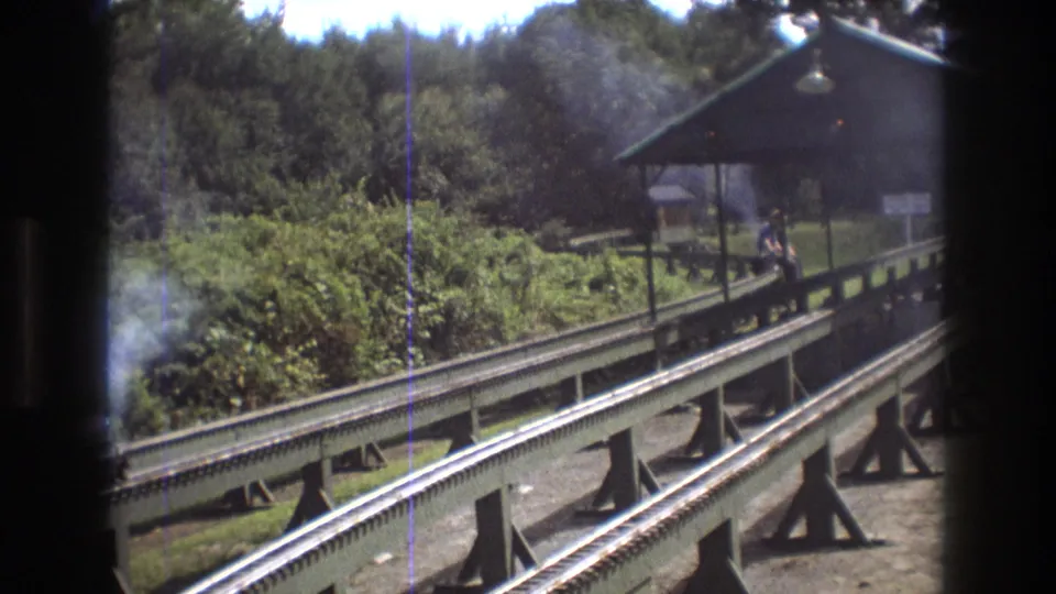 A miniature railway passes the station platform in the wooded Baxter, Maine setting, with the rider and train partly visible beyond the track.