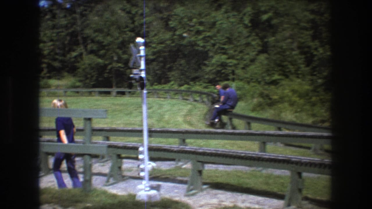 1975 Baxter, Maine 8mm frame showing people around a green miniature rail track, with a tall crossing signal in the foreground and trees behind.