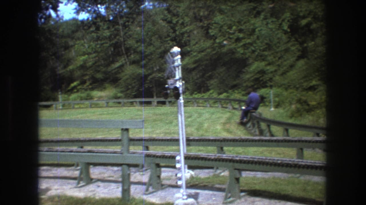 1975 Baxter, Maine 8mm frame showing a rider moving away on a miniature track near a tall crossing signal, with mowed grass and dense trees around the route.
