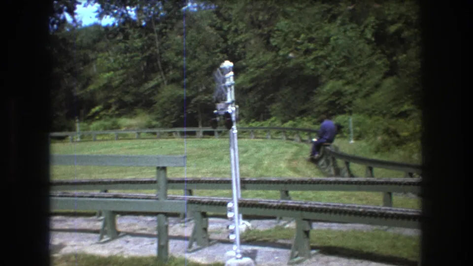 1975 Baxter, Maine 8mm frame showing a rider moving away on a miniature track near a tall crossing signal, with mowed grass and dense trees around the route.