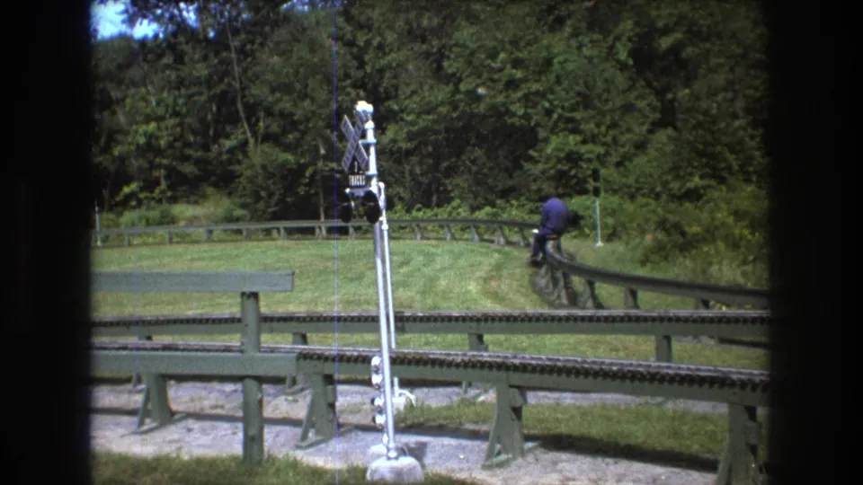 1975 Baxter, Maine 8mm frame with a crossing signal centered near the miniature rail line and a person riding along the track through a grassy wooded setting.