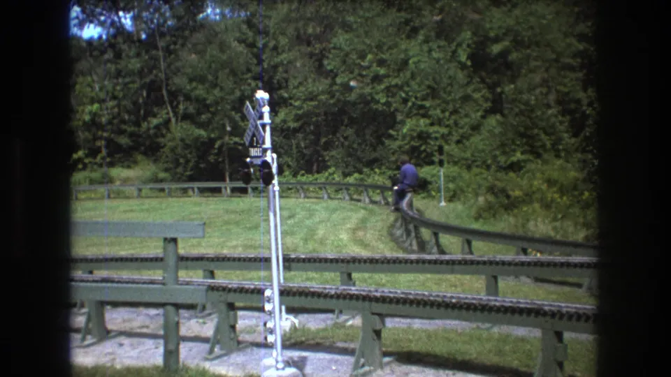 1975 Baxter, Maine 8mm frame showing a rider on a small rail vehicle or track seat moving away from the camera, with a crossing signal and wooded lawn visible.
