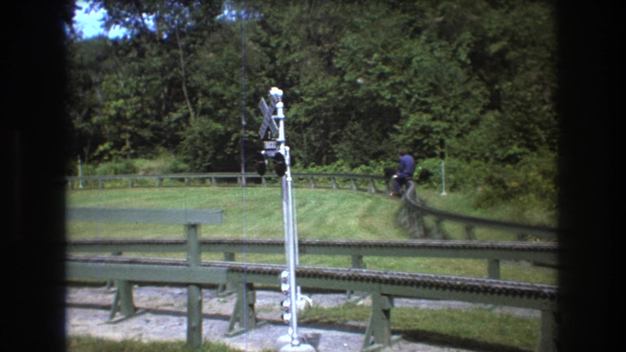 1975 Baxter, Maine home-movie frame showing a distant man in a blue shirt on a small vehicle beside a curved outdoor track, with a railroad crossing signal, grass, and trees visible.