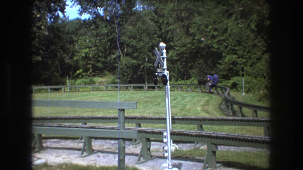 1975 Baxter, Maine frame with a man riding or sitting on a small vehicle along a curved track, seen near a railroad crossing signal and grassy wooded surroundings.