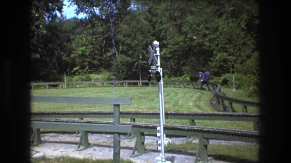 1975 Baxter, Maine frame with a man riding or sitting on a small vehicle along a curved track, seen near a railroad crossing signal and grassy wooded surroundings.