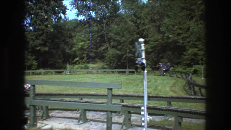 1975 Baxter, Maine home-movie frame showing a distant rider on a small track vehicle, partly obscured by foreground railings and a railroad crossing signal, with trees beyond.