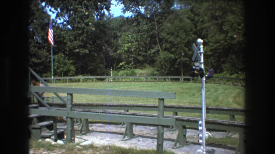1975 Baxter, Maine frame with a railroad crossing signal in the foreground, a curved outdoor track, an American flag, green railings, and a distant seated rider near the trees.