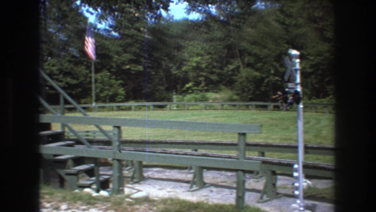 1975 Baxter, Maine 8mm frame showing a miniature train track, green wooden railings, grass, trees, and an American flag viewed from a rider-like perspective.