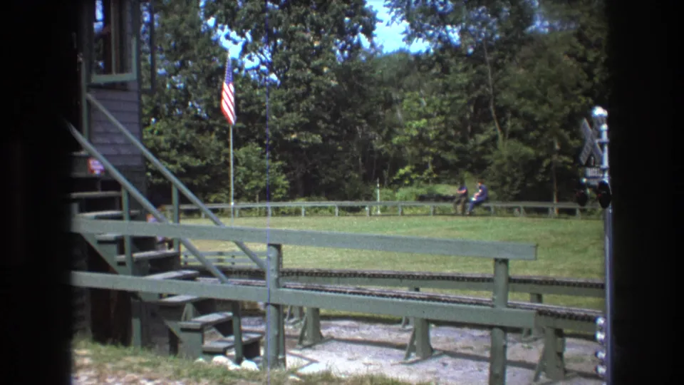 1975 Baxter, Maine 8mm view of a miniature train attraction with green wooden structures, track, flag, grassy field, trees, and distant visitors.