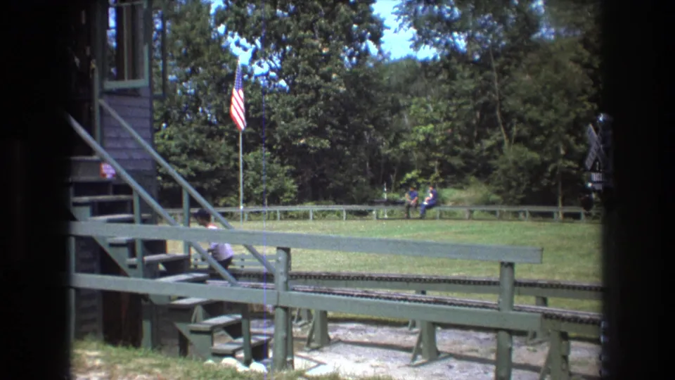 1975 Baxter, Maine frame showing the miniature railway foreground, green railings, American flag, wooded backdrop, and distant visitors crossing or sitting in the park.