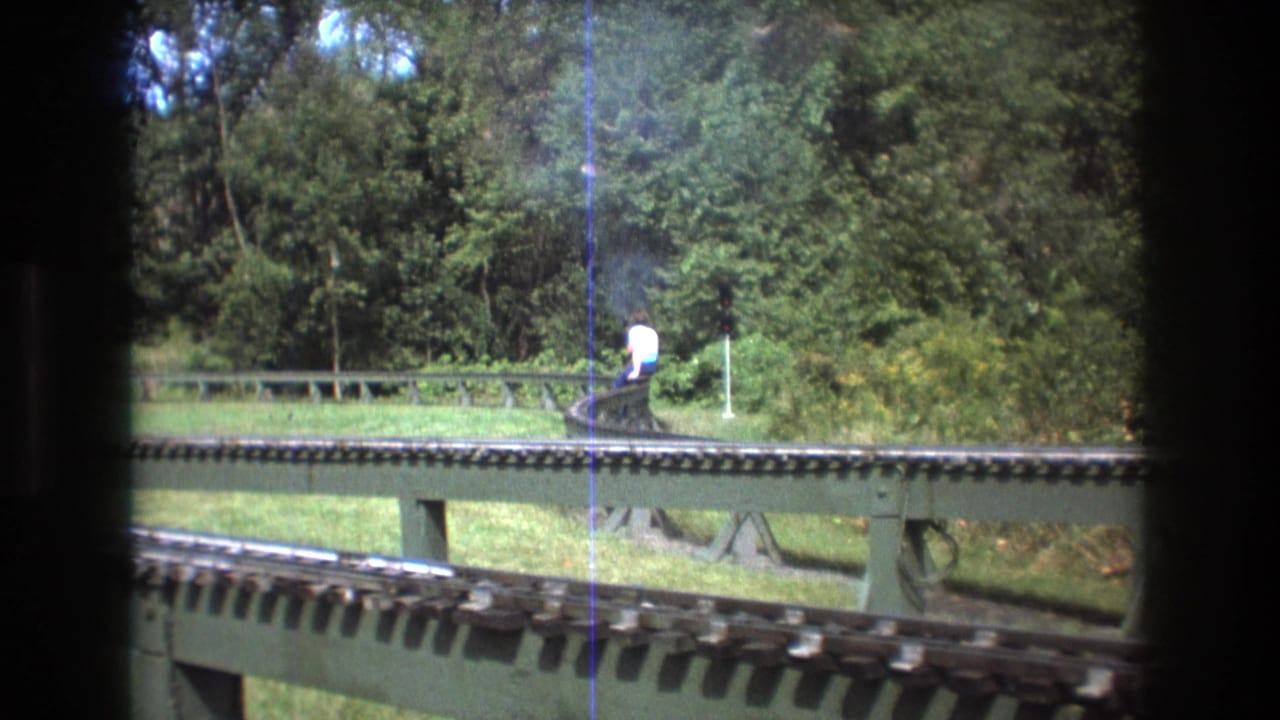 1975 Baxter, Maine 8mm frame showing elevated miniature railway tracks in a wooded park, with two distant adults riding a small train near a curve.