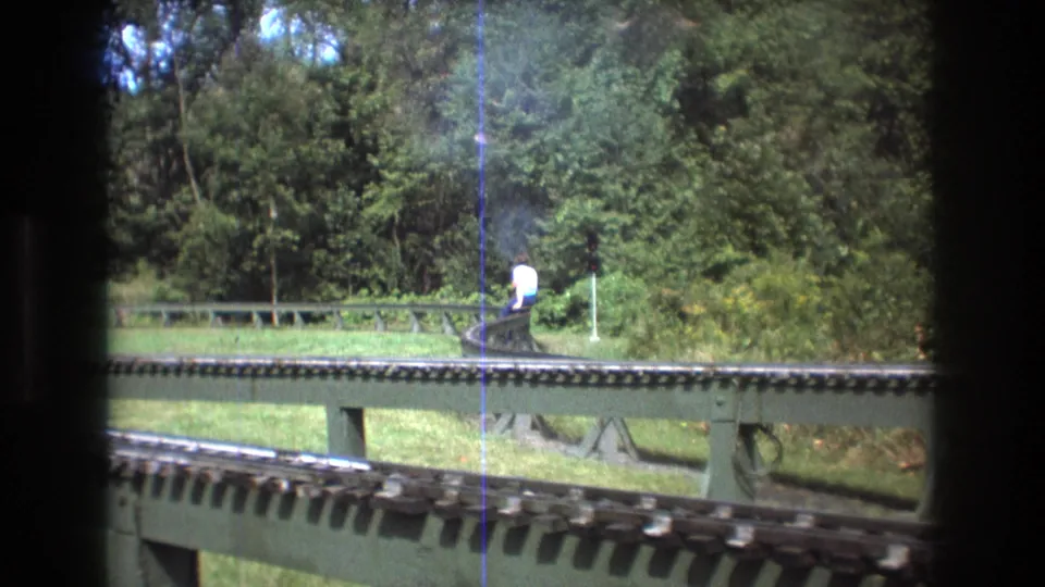 1975 Baxter, Maine 8mm frame showing elevated miniature railway tracks in a wooded park, with two distant adults riding a small train near a curve.