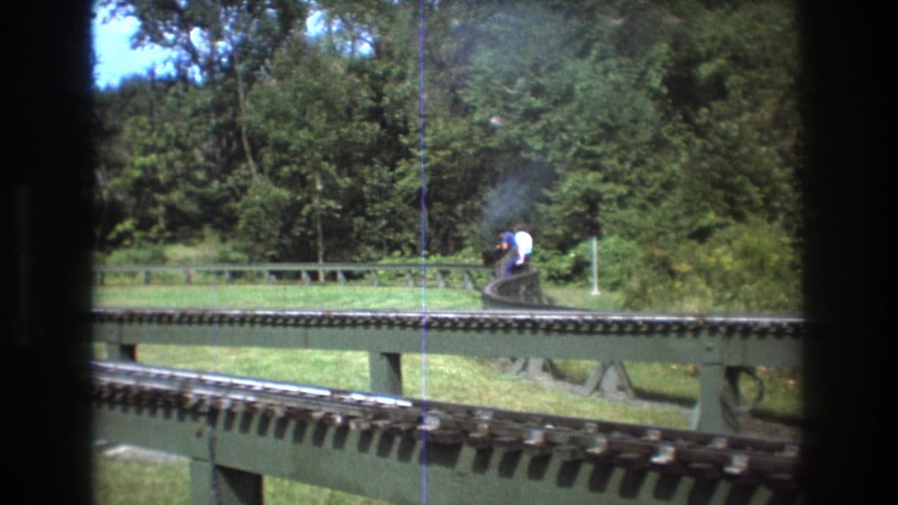 Small train with two riders moves along elevated tracks beside trees and grass.