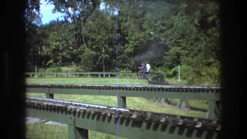 1975 Baxter, Maine 8mm frame showing a miniature train with adult riders rounding a curve on elevated rails in a wooded park setting.