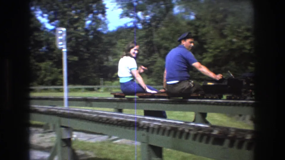 1975 Baxter, Maine close frame of an adult man in a blue shirt and cap operating a small black miniature train with an adult woman in a white top seated behind him.