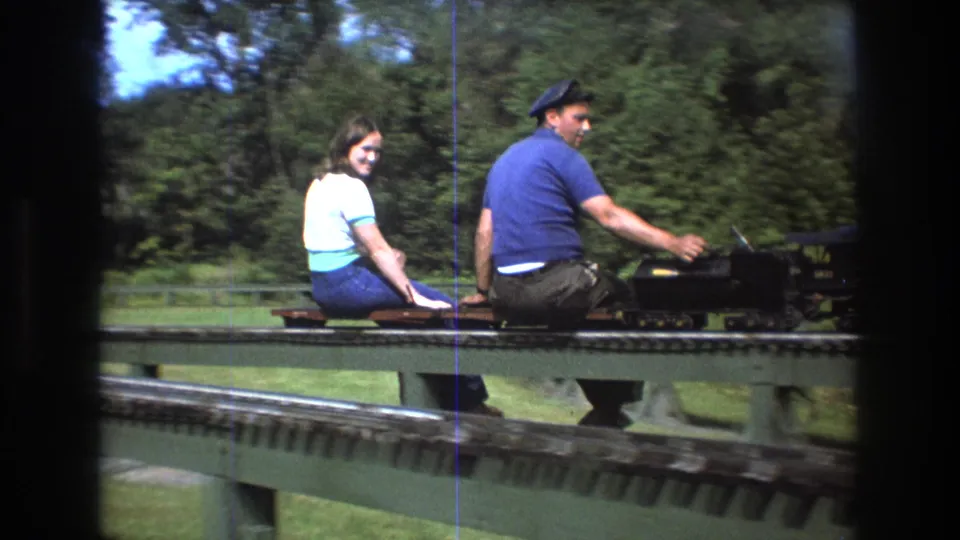 In Baxter, Maine in 1975, a young woman in a white top and jeans sits beside a young man in a blue shirt and cap on a miniature train crossing a green bridge.
