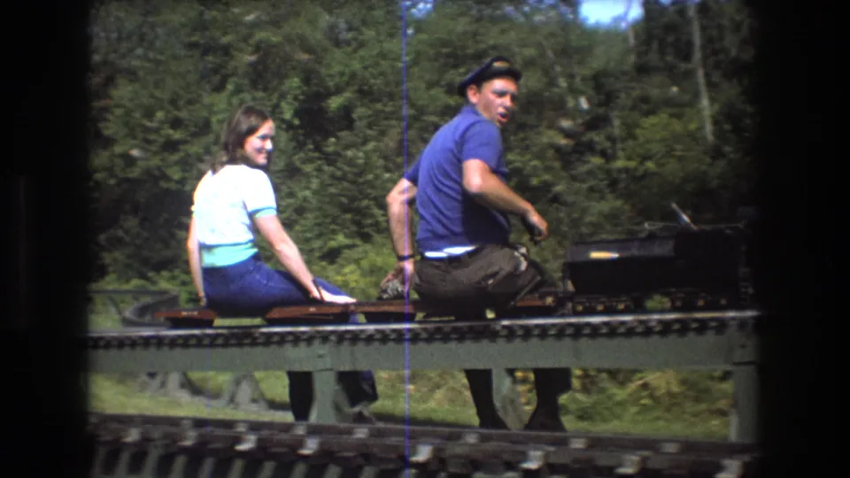 In the 1975 Baxter, Maine home-movie scene, the young woman smiles back toward the camera while the young man operates or rides the small open train.