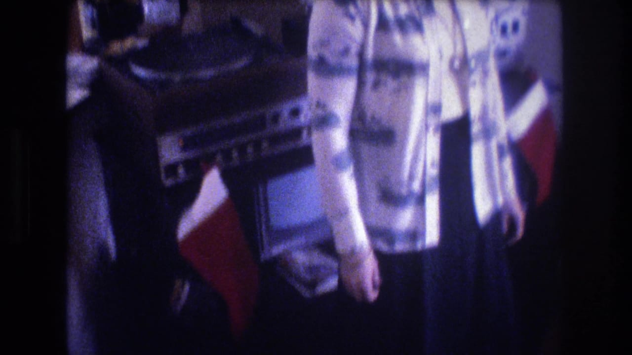 Adult woman in patterned clothing stands in a Baxter, Maine, living room near household audio or television equipment and a red Christmas stocking in 1975 home-movie footage.