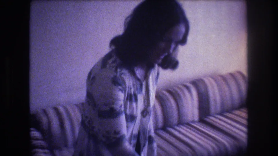 Adult woman sits on a striped sofa in a Baxter, Maine, private living room, captured in soft, blue-shifted 1975 8mm home-movie footage.