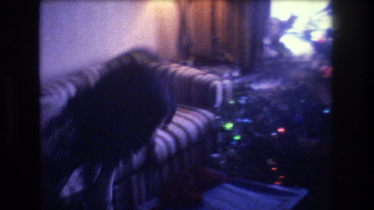 1975 Baxter, Maine 8mm frame showing a dim private living room with a striped armchair, soft Christmas tree lights, and an indistinct adult figure moving near the seating area.