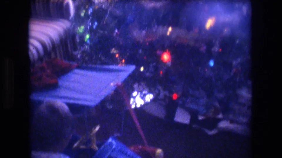 1975 Baxter, Maine 8mm frame showing blurred multicolored Christmas lights, a striped armchair, reflective ornaments, and papers or cards on a table in a private living room.