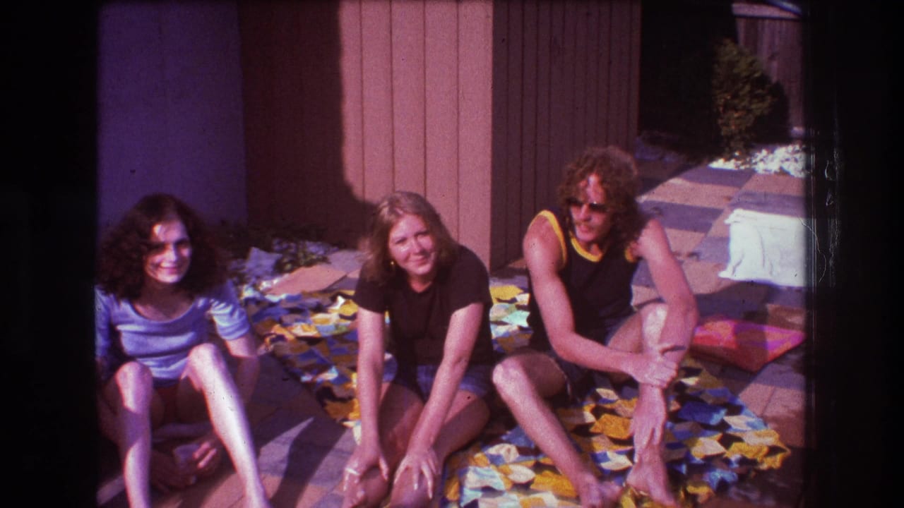 Three people sit on a colorful blanket in a bright backyard patio scene from the supplied 1975 Boston home-movie clip.