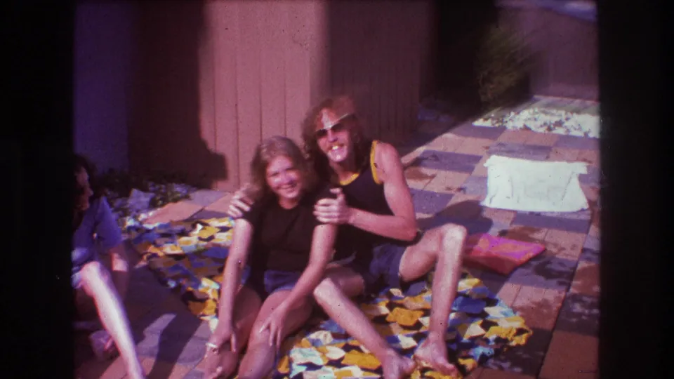 A woman and a man in sunglasses sit close together on a patterned blanket, with a third seated person partly cropped at the left edge.