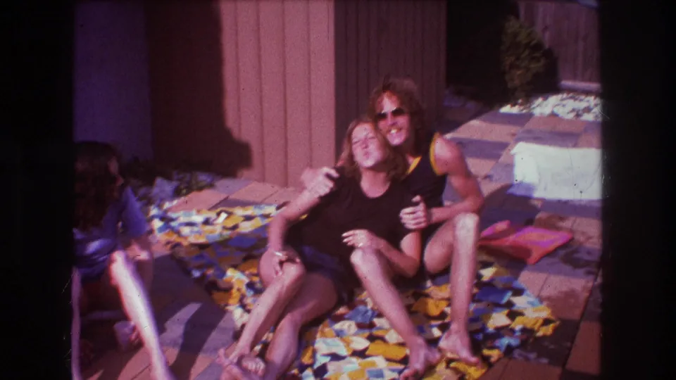 The man in sunglasses wraps an arm around the seated woman as they pose on the colorful blanket in the Boston 1975 home-movie scene.