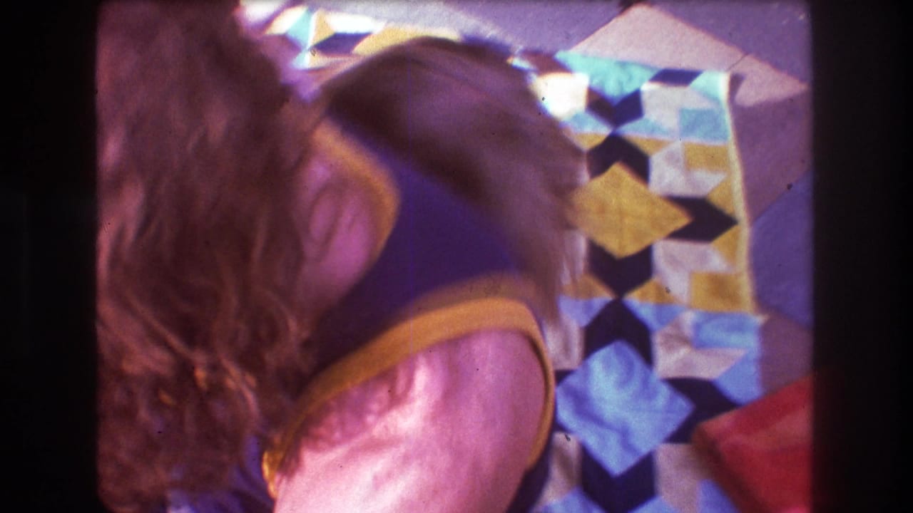 1975 Boston home-movie frame showing two people leaning together on colorful geometric blankets, with soft focus and warm 8mm color.