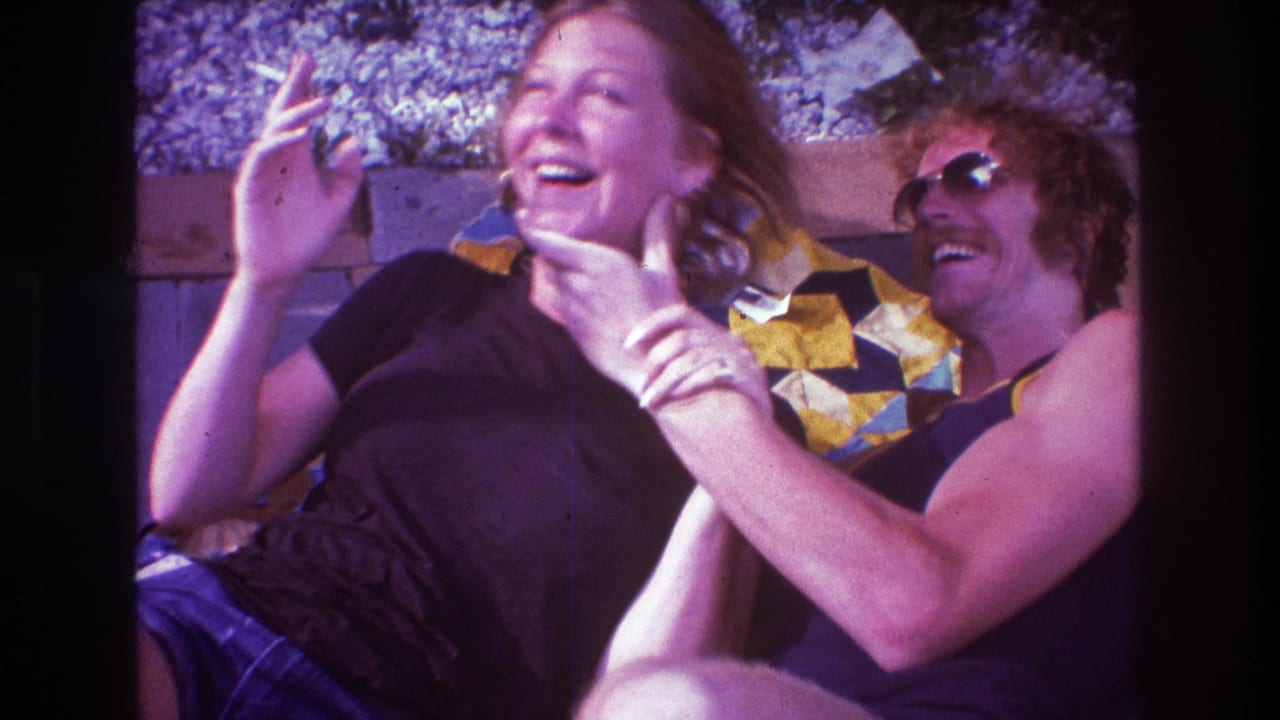 1975 Boston 8mm frame showing two adults laughing and playfully leaning together on patterned blankets in an outdoor private setting.