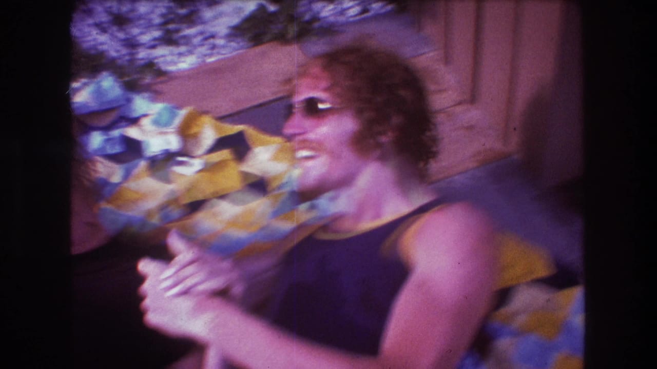 A blurred 1975 Boston home-movie frame shows an adult in sunglasses laughing while reclining near a bright patterned blanket.