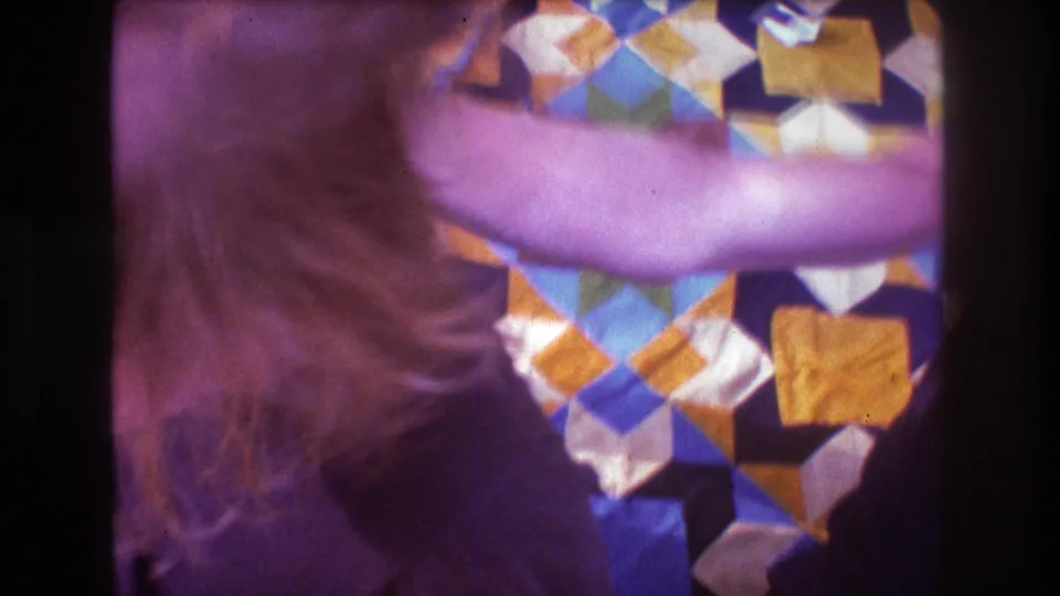Motion-blurred 8mm frame from the 1975 Boston clip showing close physical play on a patterned blanket, with hair and an arm crossing the foreground.