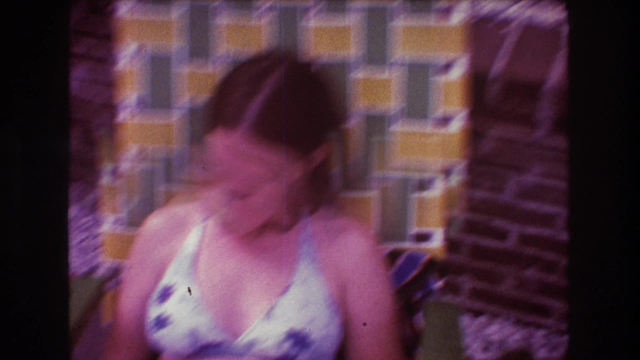 In 1975 Boston home-movie footage, a woman in a floral bikini top sits in a folding chair by a patterned yellow and green tiled wall, blurred as she turns her head.