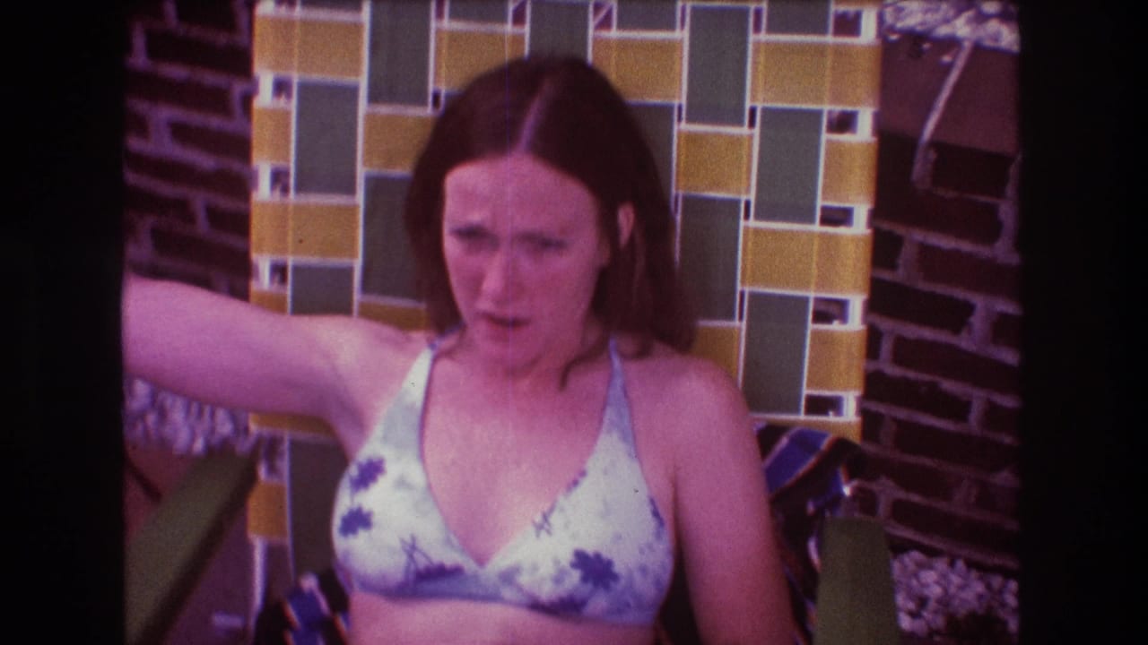 In 1975 Boston home-movie footage, a woman in a floral bikini top sits against a yellow and green tiled wall, one arm raised on the chair as she looks toward the camera.