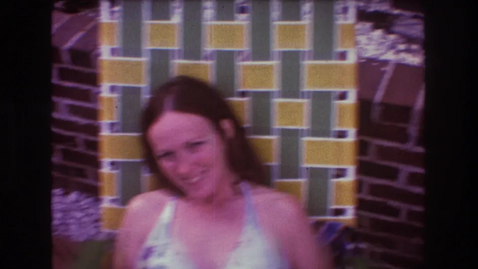 In 1975 Boston home-movie footage, the seated woman smiles toward the camera near the patterned patio wall, with soft focus and visible 8mm color grain.