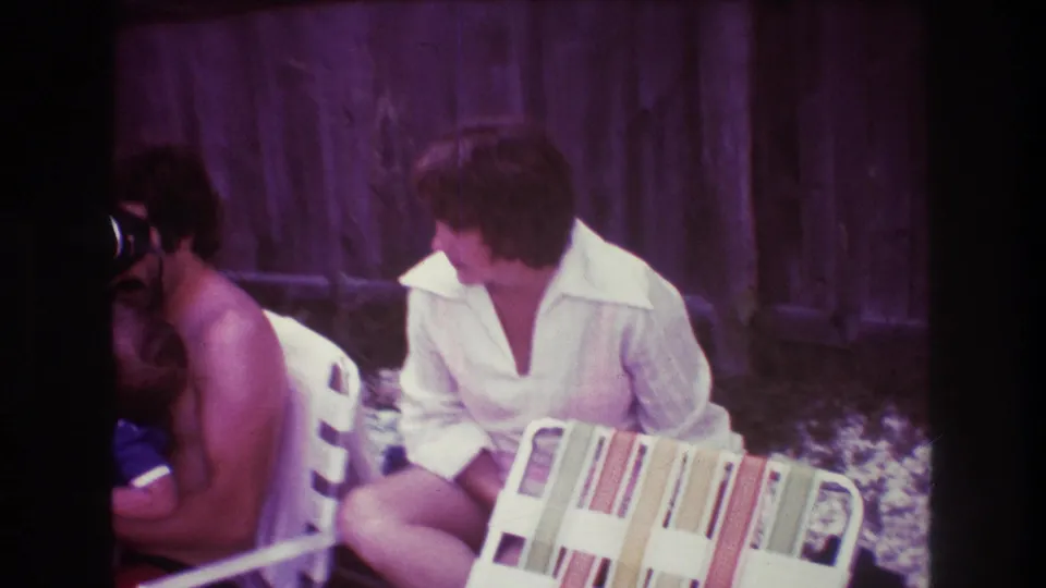 In 1975 Boston home-movie footage, two adults sit outdoors near poolside chairs, including one person in a white open-collar shirt beside a striped chair back and wooden fence.