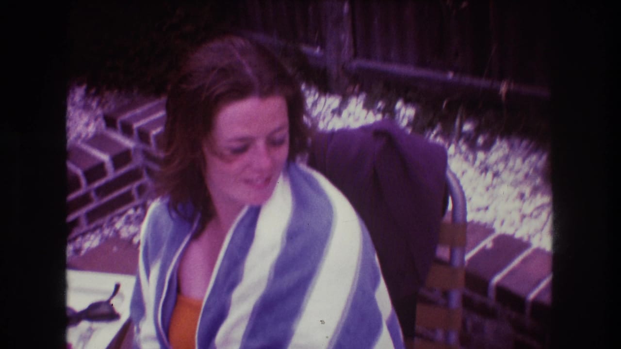 In 1975 Boston home-movie footage, a woman in an orange top sits outdoors near brickwork with a blue and white striped towel around her shoulders.