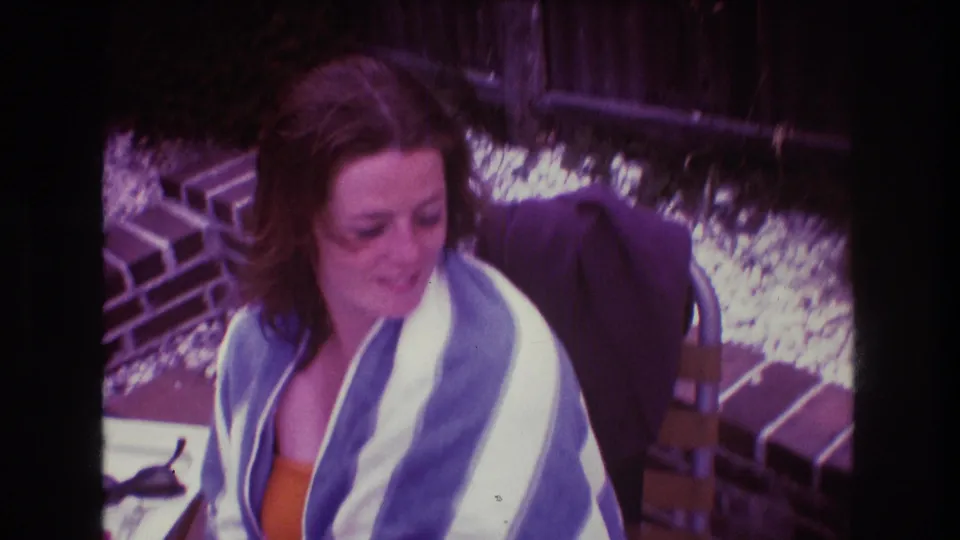 In 1975 Boston home-movie footage, a woman in an orange top sits outdoors near brickwork with a blue and white striped towel around her shoulders.