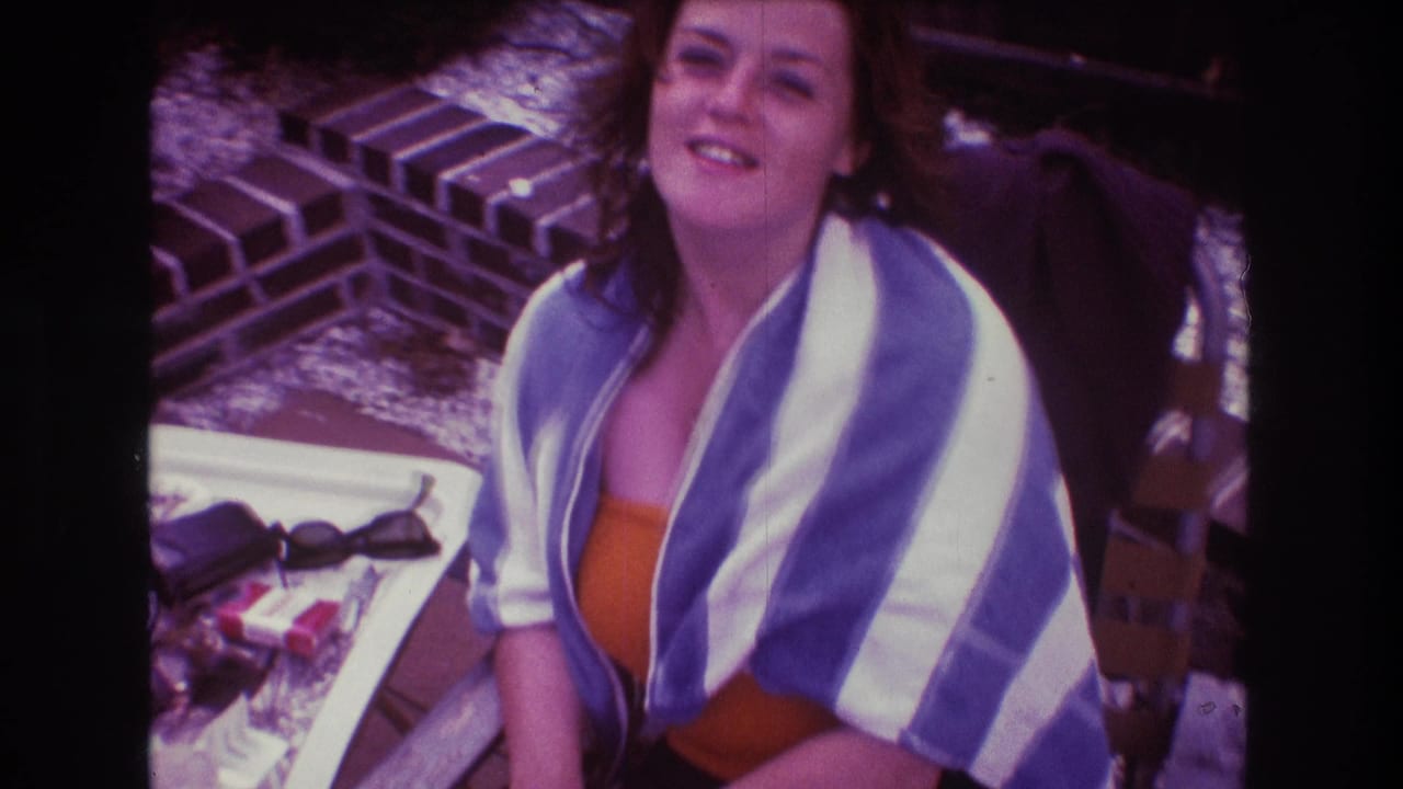 A woman seated outdoors in 1975 Boston looks toward the camera while wrapped in a blue and white striped towel, with brickwork and a small table nearby.