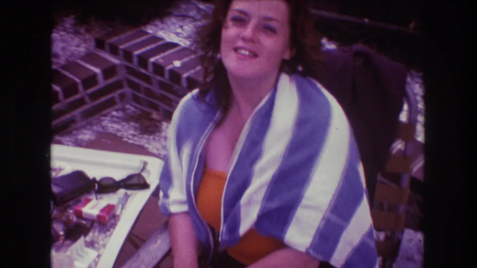 A woman seated outdoors in 1975 Boston looks toward the camera while wrapped in a blue and white striped towel, with brickwork and a small table nearby.