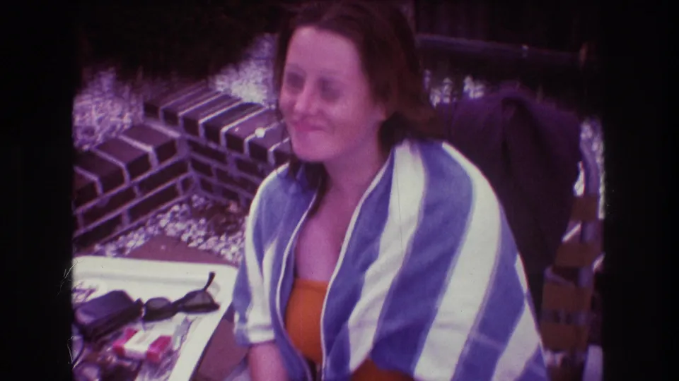 The 8mm frame shows a smiling woman in an orange top and striped towel seated near a brick outdoor fireplace and a small table.