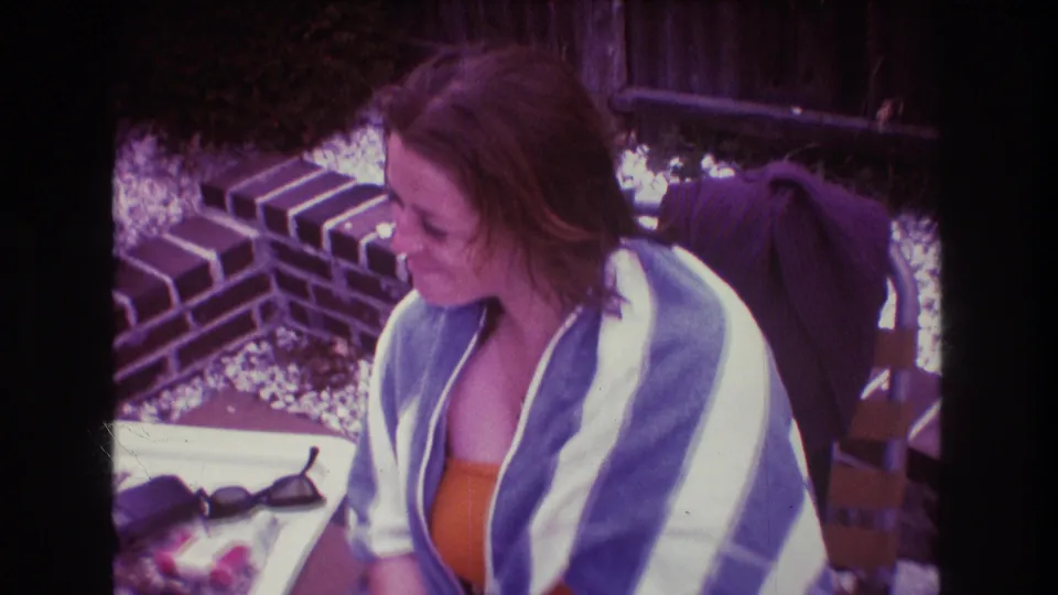 A woman wrapped in a blue and white striped towel looks down beside patio furniture, brickwork, sunglasses, and a small table in 1975 Boston home-movie footage.
