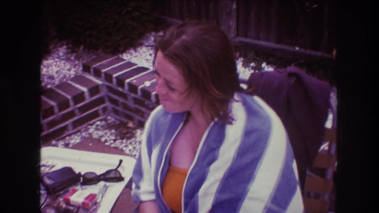 In 1975 Boston home-movie footage, an adult woman in an orange swimsuit and blue and white striped towel sits outdoors at a table near brick edging and gravel.