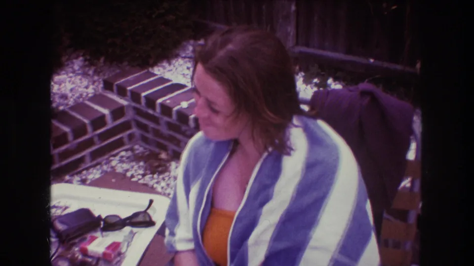 In 1975 Boston home-movie footage, an adult woman in an orange swimsuit and blue and white striped towel sits outdoors at a table near brick edging and gravel.
