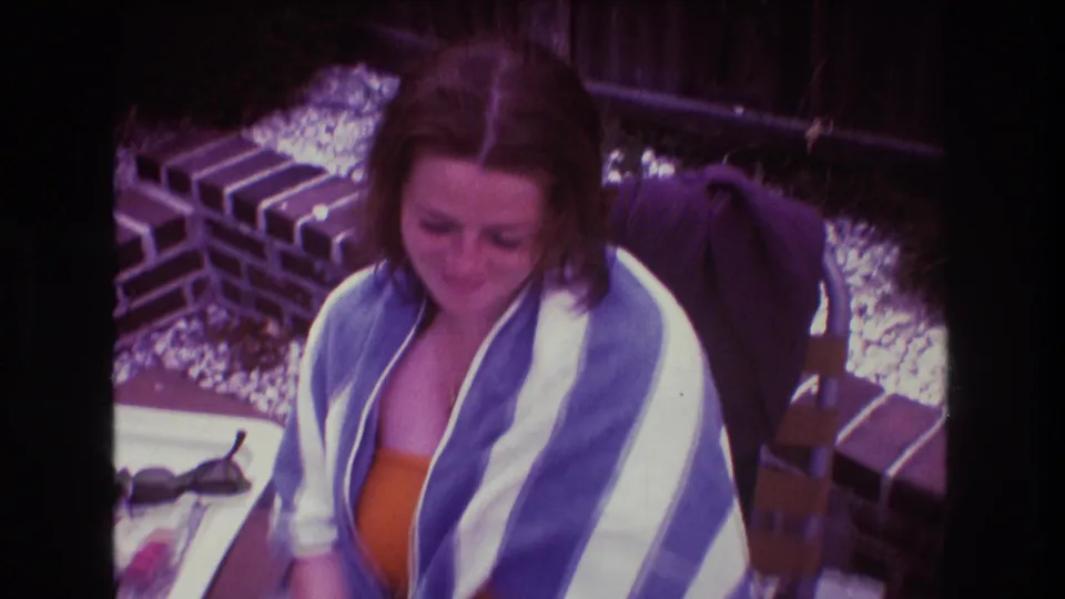 A soft 8mm frame shows the woman seated close to the outdoor table, wrapped in a striped towel with the brick border, gravel, and fence behind her.