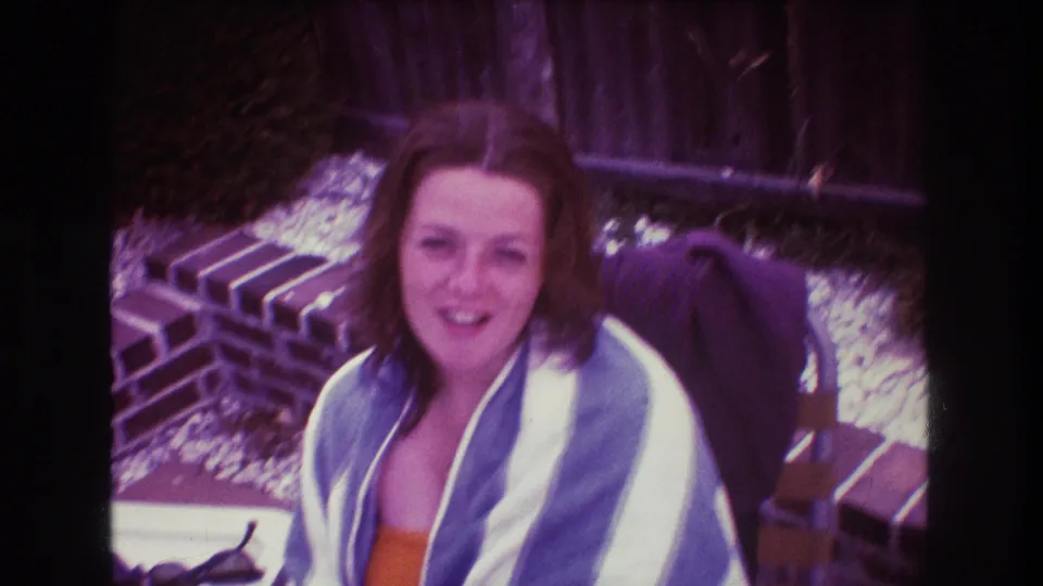 The woman smiles toward the handheld camera in the 1975 Boston home-movie frame, seated outdoors with a towel around her shoulders and patio details behind her.