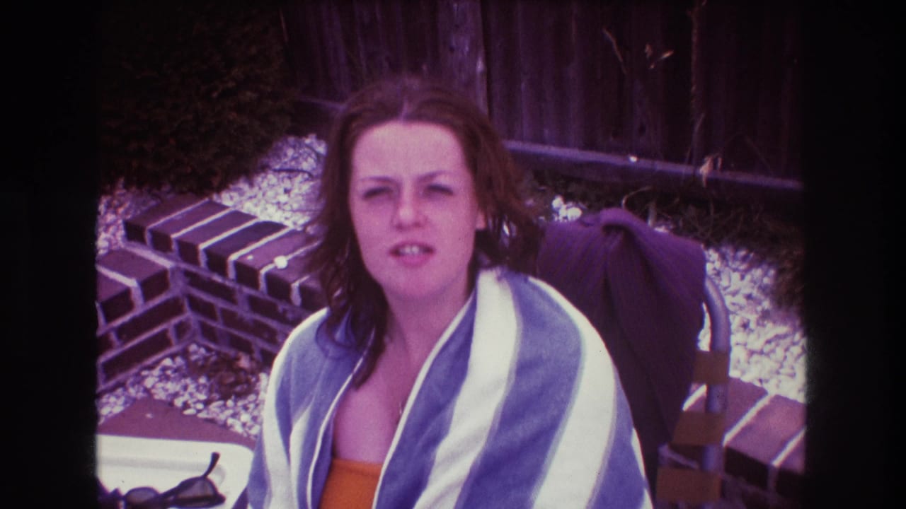 In a 1975 Boston 8mm home-movie frame, an adult woman in an orange swimsuit and blue and white striped towel looks toward the camera beside a low brick garden wall.