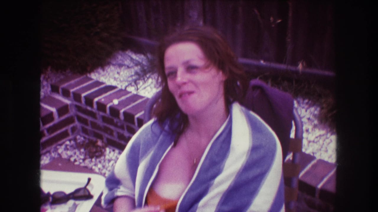 The woman remains seated outdoors in the 1975 Boston home-movie shot, wrapped in a striped towel over an orange swimsuit with a low brick wall and gravel behind her.
